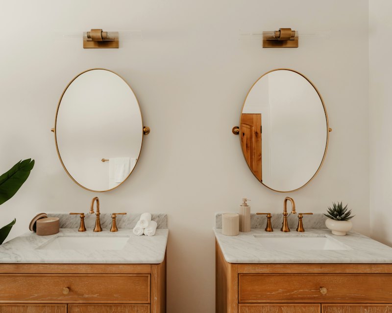 Double vanity with brass wall sconces over oval mirrors